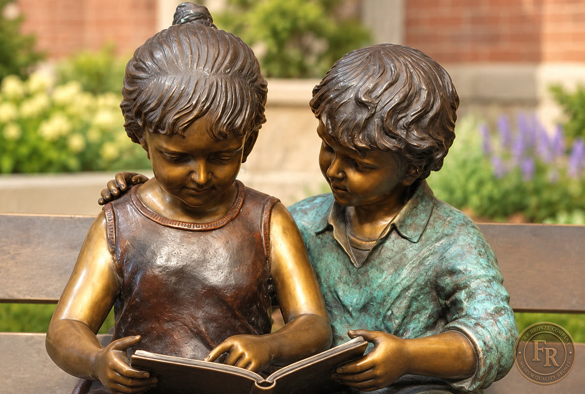 Frontal school-garden concept close-up of the bronze boy and girl reading together, with the open book and contrasting patina.