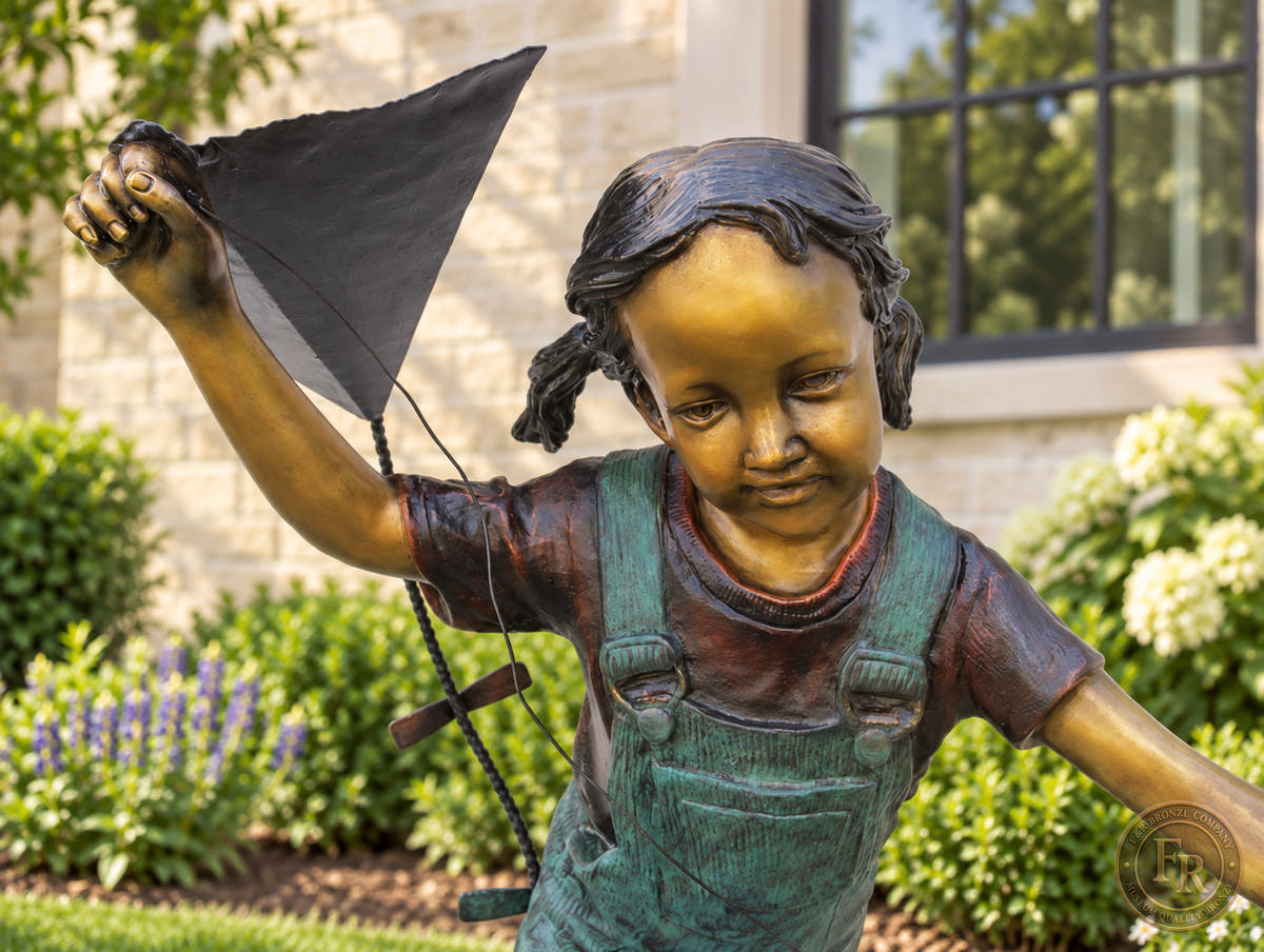 Close-up of the bronze girl’s smiling face, braided hair and raised hand holding the kite, with warm bronze and turquoise patina.