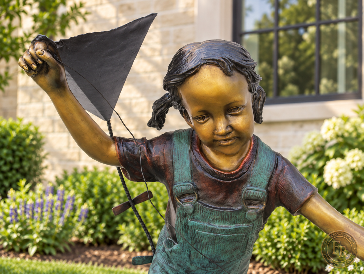 Close-up of the bronze girl’s smiling face, braided hair and raised hand holding the kite, with warm bronze and turquoise patina.